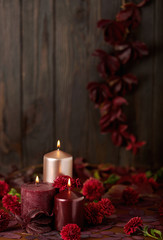 Three candles of crimson and pink color on a dark background with cones, leaves and daisies.