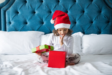 Pretty little girl is surprised while opening a gift box and smiling while sitting on her bed in her room at home