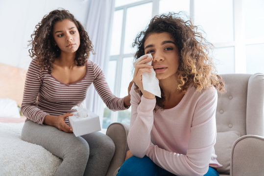 Sad Times. Selective Focus On An Emotional Girl Wiping Her Eyes With A Tissue While Crying Next To Her Careful Sibling Calming Her Down.