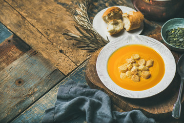 Fall warming pumpkin cream soup with croutons and seeds on board over rustic wooden background, copy space, selective focus. Autumn vegetarian, vegan, healthy comfort food concept