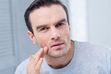 Strange rush. Concerned earnest unhappy man looking at the camera and scrutinizing his face while touching face with a finger