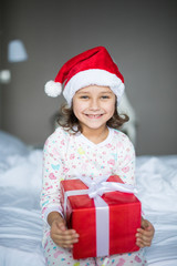 Charming kid girl in Santa's hat looking at camera with smile with her gift box while sitting in white bed