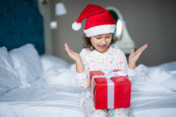 Surprised little girl in Santa's hat holding present while sitting with crossed legs in morning bed