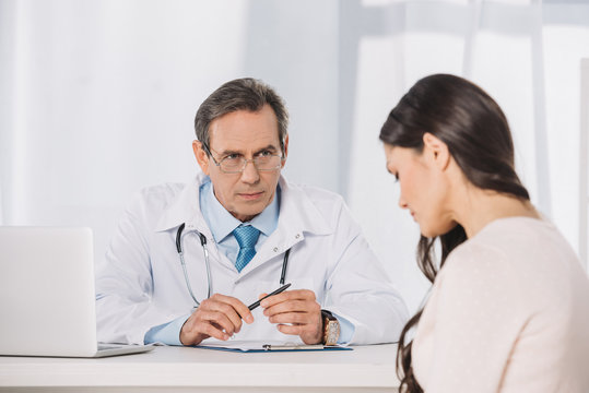 Male Serious Doctor And Female Patient Sitting At Talking At Clinic