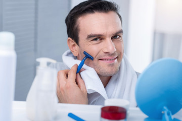 Promising day. Merry cheerful young man shaving in the morning and touching his chick with a towel while carrying razor 