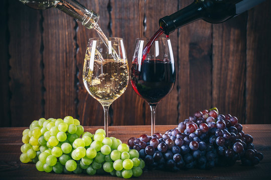 Fototapeta Pouring red and white wine into the  wine glasses with a bunch of red and white grapes against wooden background
