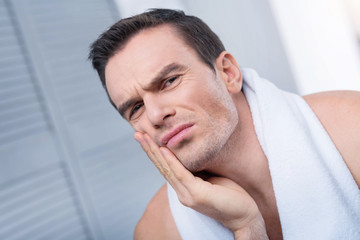 Obraz premium Horrible toothache. Attractive sad pleasant man pressing hand against the chick and staring at the camera while towel hanging on his shoulders 