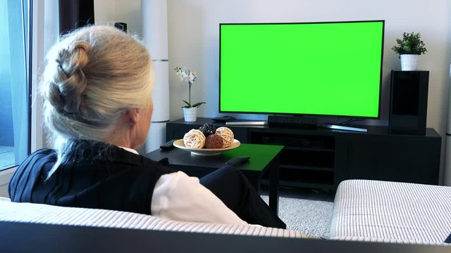 Old caucasian woman watches television in living room - green screen