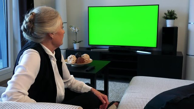Old caucasian woman watches television in living room and then talks to camera