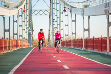 Fototapeta premium Two Young Female Cyclists Riding Road Bicycles on Bridge Bike Line in Cold Sunny Autumn Day. Healthy Lifestyle Concept.