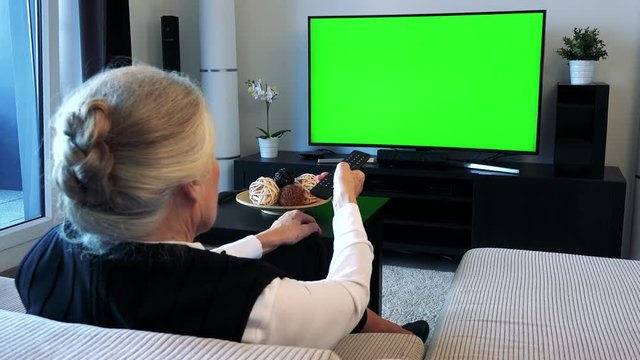 Old caucasian woman watches television in living room and switches program 