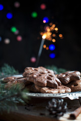 gingerbread cookies for the new year on a silver tray, New Year's lights, fireworks, fur-tree, cones, cinnamon on a wooden cutting board on a dark background