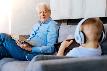 Family evening. Happy nice cheerful man sitting together with his grandson and looking at his while reading a book