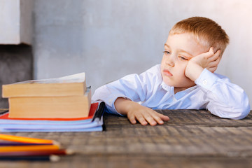 Naklejka premium Time for homework. Unhappy nice serious boy sitting at the desk and looking at his textbooks while preparing to do his homework