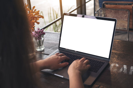 Mockup image of hands using and typing on laptop with blank white screen and flower vase on vintage wooden table in cafe
