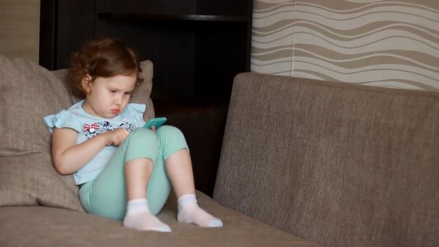 Funny child looks at the phone screen and plays downloaded application on a smart phone close-up. A little cute girl lies in sofa in a living room, looking cartoon and playing the game