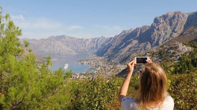 Woman with smartphone makes a photo of sea bay among mountains, seaside town and cruise liner