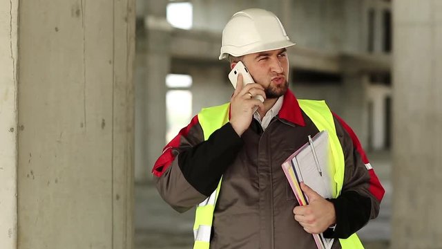 Engineer in white helmet talks on smartphone at construction site. Builder in green vest talks on telephone on project site. Worker holds in hands works papers and cell phone
