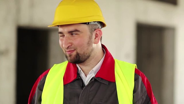 Funny builder on construction site make faces. Worker in hard hat stands on project site, looks at the camera and makes grimaces