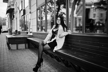 Obraz premium Black hair sexy woman in glasses and coat sitting on bench with mobile phone at hand.