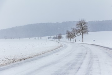 Fototapeta na wymiar Eisige glatte Winter Straße mit Schnee wartet auf Winterdienst. Die Fahrbahn ist weiß glatt verschneit. Vorsicht vor Unfall durch Schneesturm und schlechte Sicht. Wetter zur Weihnachten Jahreszeit