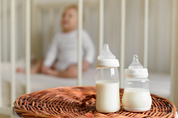 Bottle with milk for baby on a straw basket. Baby in a kids room with a bed on a background. - 183626377