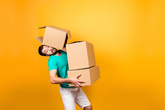 It's moving day! Busy funny guy in casual clothes is carrying big boxes in his hands and trying to keep one using his head, isolated on yellow background