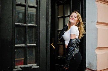 Elegant blonde girl wear on black leather jacket posing at streets of town background old doors.