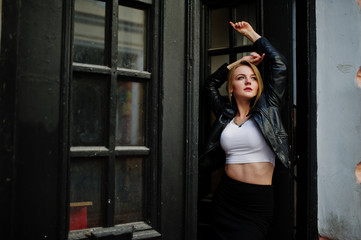 Elegant blonde girl wear on black leather jacket posing at streets of town background old doors.