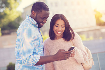 Interesting shot. Delighted positive afro american man standing near his girlfriend and holding a smartphone while showing her a picture on it