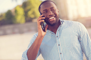 In touch. Happy pleasant afro american man smiling and talking on the phone while staying in touch with his friends
