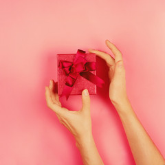Woman's hands reaching for a present in red box on pink background, romantic flat lay, toned image, square