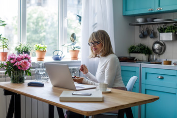 Pretty senior Caucasian woman eating dessert and reading on laptop at her home.