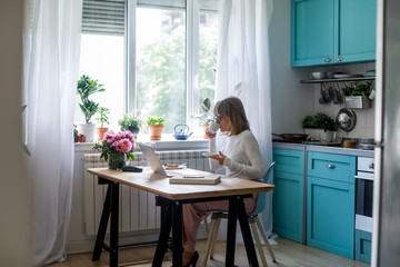 Pretty senior Caucasian woman drinking coffee and reading on laptop at her home.