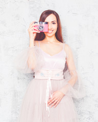 Stunning young girl in a chic pink airy dress, smiling and taking a picture with a vintage pink camera.