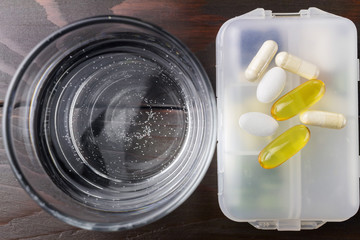 White and yellow capsules of glucosamine, omega-3 pills of calcium on plastic box, glass with water at wooden table, top view