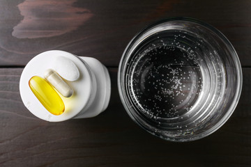 Complex of vitamins and dietary supplement, yellow capsule of omega 3, white capsule of glucosamine, calcium pill on the white plastic bottle and glass of water at wooden table, top view