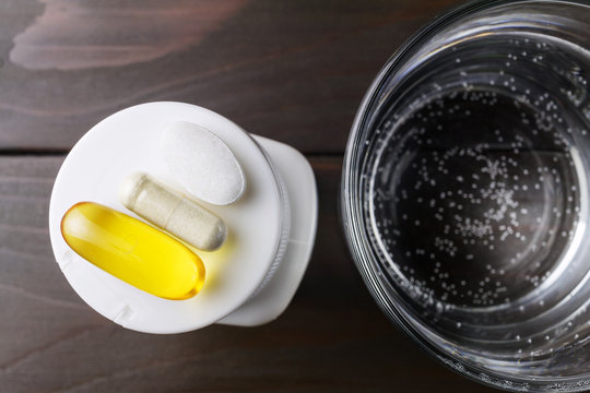 Complex of vitamins and dietary supplement, yellow capsule of omega 3, white capsule of glucosamine, calcium pill on the white plastic bottle and glass of water at wooden table, top view