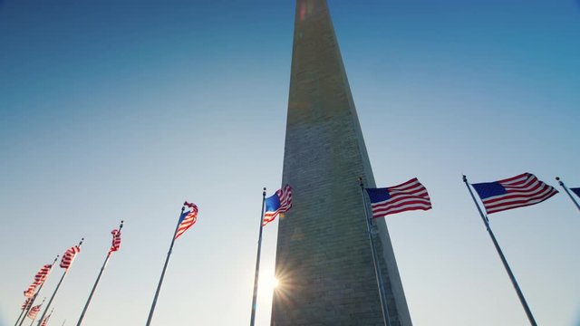 Tilt shot: The sun appears because of the Washington monument. American flags flap below