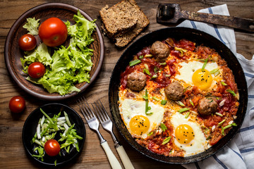 shakshuka, the Israeli dish of eggs, tomatoes and meatballs with greens on wooden background