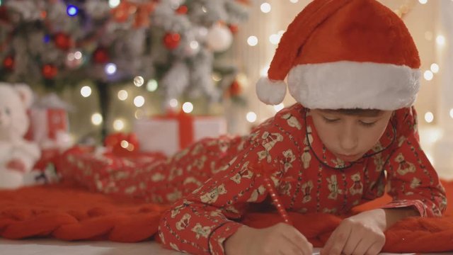 Boy in Santa's cap and pajamas lies on the knitted plaid and writes the letter to Santa