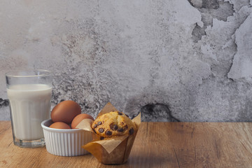 Muffin with milk chocolate, eggs in a white bowl and a glass of almond milk on a brown wooden table against the background of an empty concrete wall