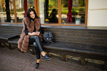 Fototapeta na wymiar Fashion outdoor photo of gorgeous sensual woman with dark hair in elegant clothes and luxurious sleeveless fur coat and with backpack sitting on bench with mobile phone at autumn city.