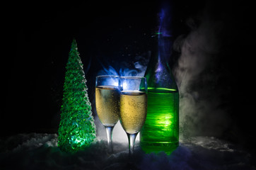 New Years Eve celebration background with pair of flutes and bottle of champagne with christmas tree on snow on dark background.