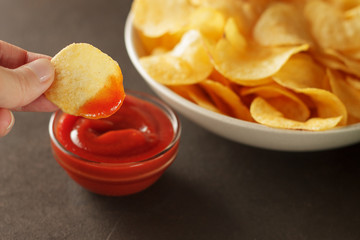 Hand dips potato chip into tomato sauce in glass bowl.