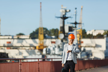 Handsome young unshaven successful business man in gray suit and protective construction orange helmet holding case, looking time on watch, walking in sea port against cargo ship and crane background