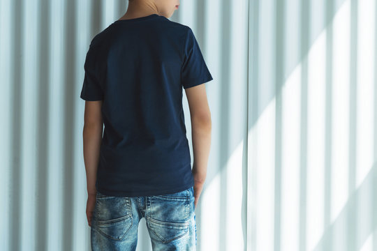 Back view. Young hipster boy dressed in black t-shirt is stands indoor against white wall. Mock up. Space for logo, text, image.