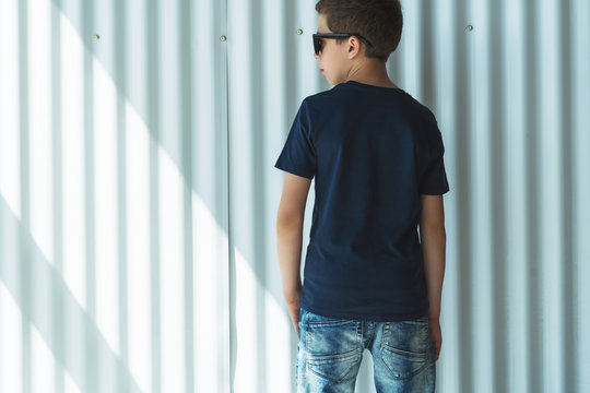 Back view. Young hipster boy dressed in black t-shirt is stands indoor against white wall. Mock up. Space for logo, text, image.