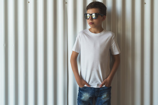 Front view. Young hipster boy in sunglasses dressed in white t-shirt is stands indoor against white wall. Mock up. Space for logo, text, image.