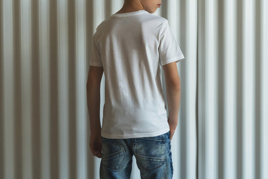 Back view. Young hipster boy dressed in white t-shirt is stands indoor against white wall. Mock up. Space for logo, text, image.